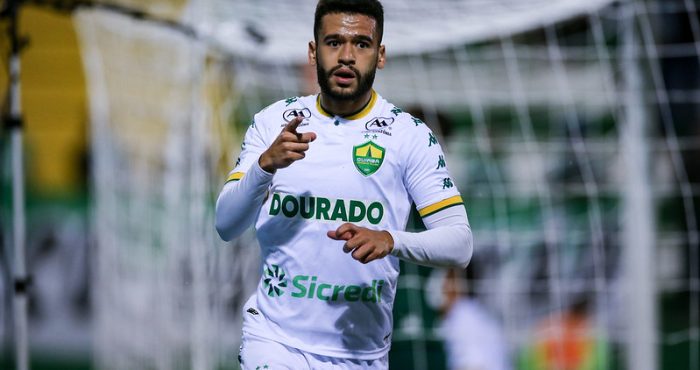 "Patrick de Lucca, volante do Cuiabá, celebra após marcar, apontando com a mão direita. Ele usa o uniforme de jogo branco, em frente a uma trave com rede no fundo."