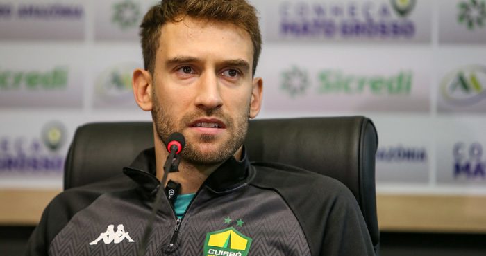 "Alan Empereur, zagueiro e capitão do Cuiabá, em coletiva de imprensa usando agasalho oficial do clube e falando ao microfone."