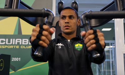 "Fotografia em plano médio de Bruno Alves, jogador do Cuiabá, realizando um exercício de supino em uma máquina de musculação. Ele é um homem pardo, de cabelos bem curtos, com expressão de foco e concentração. Veste um agasalho de treino preto com o escudo do clube e o número 3 no peito. Suas mãos usam faixas protetoras pretas enquanto seguram as alavancas do equipamento. Ao fundo, uma parede verde com o logotipo do Cuiabá Esporte Clube."