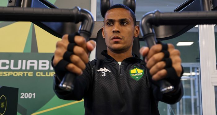 "Fotografia em plano médio de Bruno Alves, jogador do Cuiabá, realizando um exercício de supino em uma máquina de musculação. Ele é um homem pardo, de cabelos bem curtos, com expressão de foco e concentração. Veste um agasalho de treino preto com o escudo do clube e o número 3 no peito. Suas mãos usam faixas protetoras pretas enquanto seguram as alavancas do equipamento. Ao fundo, uma parede verde com o logotipo do Cuiabá Esporte Clube."