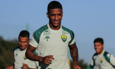 Max aparece em destaque correndo no campo durante o dia. Ele veste o uniforme de treino verde claro com o número 8 pequeno no peito. Ao fundo, outros jogadores aparecem desfocados sob um céu limpo.