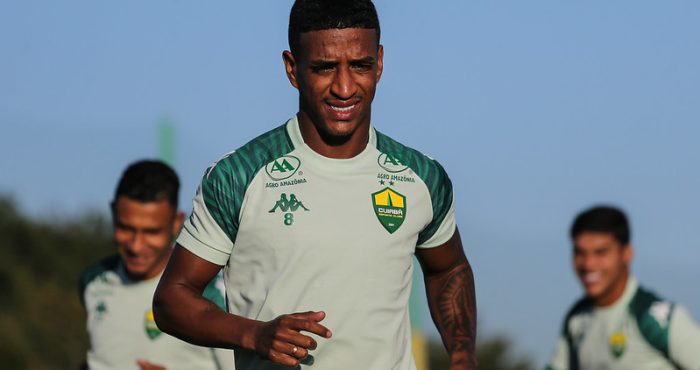 Max aparece em destaque correndo no campo durante o dia. Ele veste o uniforme de treino verde claro com o número 8 pequeno no peito. Ao fundo, outros jogadores aparecem desfocados sob um céu limpo.