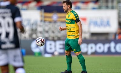 Alan Empereur, um jogador de futebol do Cuiabá, está no campo em seu uniforme verde e amarelo. Ele olha para uma bola de futebol que está no ar, perto dele. Há outro jogador com uma camisa escura no lado esquerdo da imagem, ligeiramente fora de foco.
