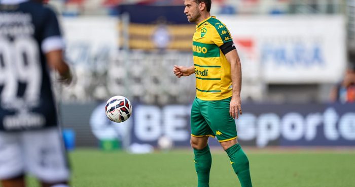 Alan Empereur, um jogador de futebol do Cuiabá, está no campo em seu uniforme verde e amarelo. Ele olha para uma bola de futebol que está no ar, perto dele. Há outro jogador com uma camisa escura no lado esquerdo da imagem, ligeiramente fora de foco.