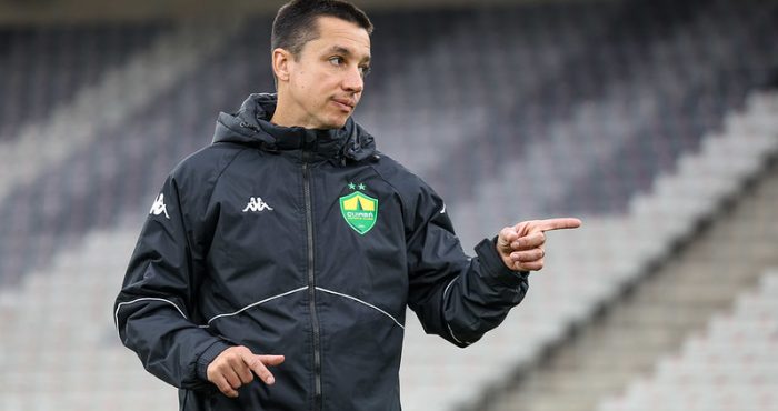 Técnico Eduardo Barros, com um agasalho preto do Cuiabá, em um estádio, gesticulando com a mão direita e apontando para frente ou para o lado, possivelmente dando instruções.