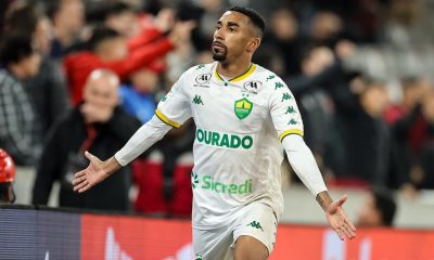 O lateral Matheusinho, do Cuiabá, celebrando em campo, vestindo a camisa branca do time. Ele está com os braços abertos e olhando para cima, em frente à arquibancada com torcedores ao fundo.