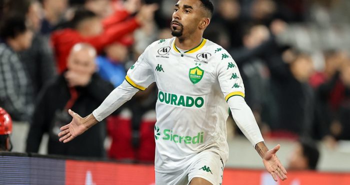 O lateral Matheusinho, do Cuiabá, celebrando em campo, vestindo a camisa branca do time. Ele está com os braços abertos e olhando para cima, em frente à arquibancada com torcedores ao fundo.