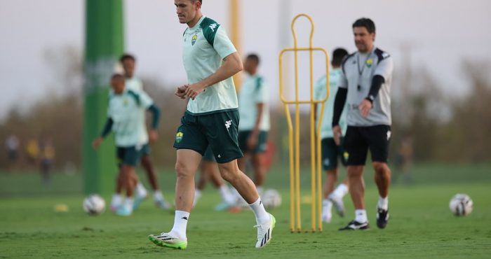 “Jogador do Cuiabá participando de atividade com bola no campo de treino, cones e boneco de treinamento ao fundo, acompanhado pela comissão técnica.”