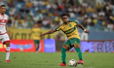 Jogador Max, do Cuiabá, em um momento de ação no campo, preparando-se para um chute ou passe longo com a perna direita. Ele está com o corpo levemente inclinado para o lado, braços abertos para equilíbrio e olhos fixos na bola. Ao fundo, é possível ver as arquibancadas com torcedores e um jogador do time adversário vestindo uniforme branco e vermelho.