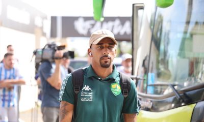 Atleta do Cuiabá chegando para o jogo, vestindo camisa polo verde do clube, boné bege e mochila, em frente ao ônibus da equipe.