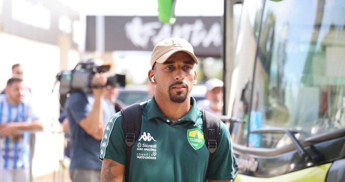 Atleta do Cuiabá chegando para o jogo, vestindo camisa polo verde do clube, boné bege e mochila, em frente ao ônibus da equipe.