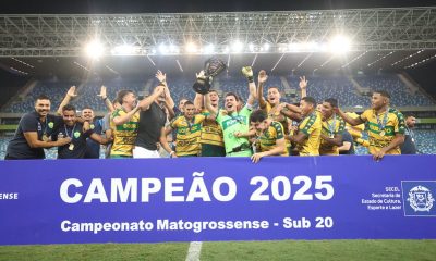 Jogadores do Cuiabá Sub-20 erguendo a taça de campeão 2025 do Campeonato Mato-grossense, sorrindo e comemorando atrás de uma faixa azul que destaca o título, dentro da Arena Pantanal iluminada.