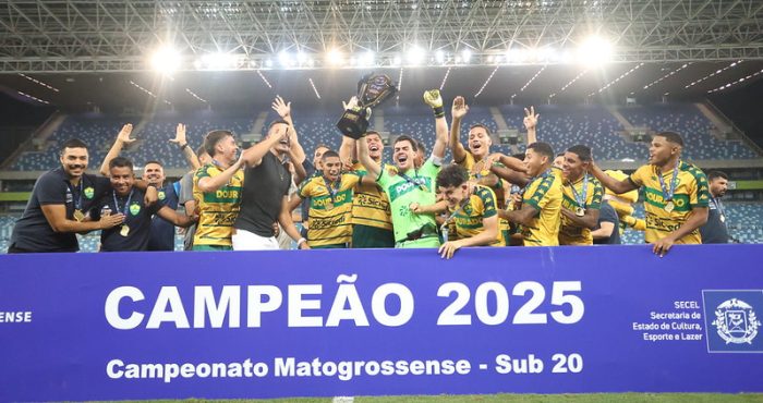 Jogadores do Cuiabá Sub-20 erguendo a taça de campeão 2025 do Campeonato Mato-grossense, sorrindo e comemorando atrás de uma faixa azul que destaca o título, dentro da Arena Pantanal iluminada.
