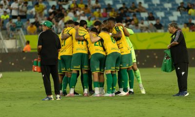 Um grupo de jogadores de futebol com uniformes amarelos e verdes, e um treinador, se reúnem em campo, possivelmente durante uma discussão tática ou celebração. O treinador, um homem mais velho com óculos e boné, está à esquerda, olhando para os jogadores que estão aglomerados no centro.