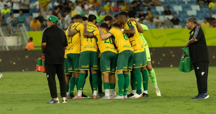 Um grupo de jogadores de futebol com uniformes amarelos e verdes, e um treinador, se reúnem em campo, possivelmente durante uma discussão tática ou celebração. O treinador, um homem mais velho com óculos e boné, está à esquerda, olhando para os jogadores que estão aglomerados no centro.