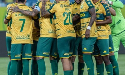Um grupo de jogadores de futebol com uniformes amarelos e verdes, e um treinador, se reúnem em campo, possivelmente durante uma discussão tática ou celebração. O treinador, um homem mais velho com óculos e boné, está à esquerda, olhando para os jogadores que estão aglomerados no centro.