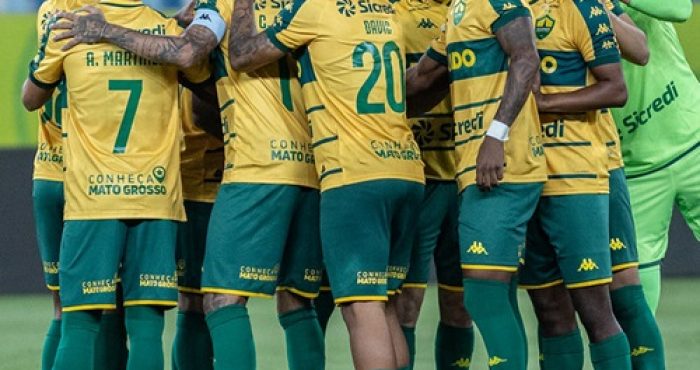 Um grupo de jogadores de futebol com uniformes amarelos e verdes, e um treinador, se reúnem em campo, possivelmente durante uma discussão tática ou celebração. O treinador, um homem mais velho com óculos e boné, está à esquerda, olhando para os jogadores que estão aglomerados no centro.