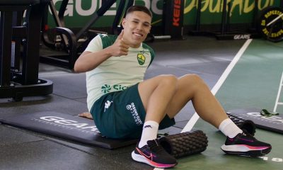 O jogador de futebol Caleb, do Cuiabá EC, está sentado em um colchonete preto de academia, sorrindo e fazendo um sinal de "positivo" com o polegar. Ele veste o uniforme de treino do clube, composto por uma camiseta verde-clara com detalhes escuros e shorts verde-escuro com a inscrição "Conheça Mato Grosso". O atleta calça tênis esportivos pretos com detalhes em rosa e branco. Ao seu lado, no chão de uma academia moderna, há um rolo de liberação miofascial e equipamentos de musculação ao fundo.