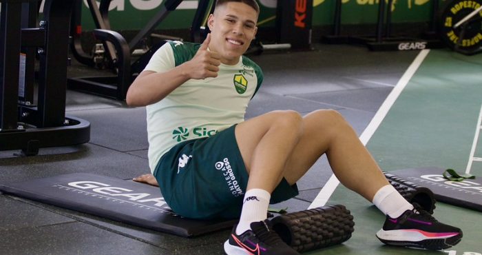 O jogador de futebol Caleb, do Cuiabá EC, está sentado em um colchonete preto de academia, sorrindo e fazendo um sinal de "positivo" com o polegar. Ele veste o uniforme de treino do clube, composto por uma camiseta verde-clara com detalhes escuros e shorts verde-escuro com a inscrição "Conheça Mato Grosso". O atleta calça tênis esportivos pretos com detalhes em rosa e branco. Ao seu lado, no chão de uma academia moderna, há um rolo de liberação miofascial e equipamentos de musculação ao fundo.