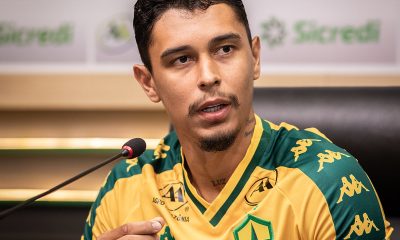 Um close-up de Vitor Mendes sentado durante uma entrevista coletiva. Ele está vestindo o uniforme de treino ou viagem do clube, que é amarelo com o escudo do Cuiabá Esporte Clube no peito e logotipos da marca Kappa nos ombros.