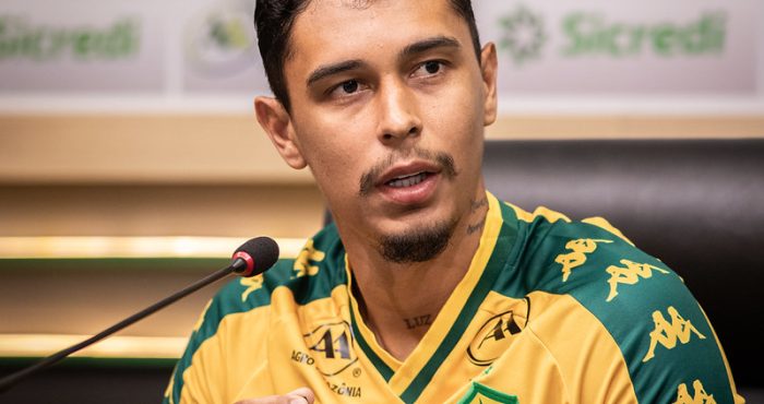 Um close-up de Vitor Mendes sentado durante uma entrevista coletiva. Ele está vestindo o uniforme de treino ou viagem do clube, que é amarelo com o escudo do Cuiabá Esporte Clube no peito e logotipos da marca Kappa nos ombros.