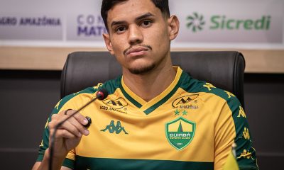 Close-up frontal de Edson sentado em uma cadeira de couro preta, posicionado atrás de um microfone durante uma coletiva de imprensa.