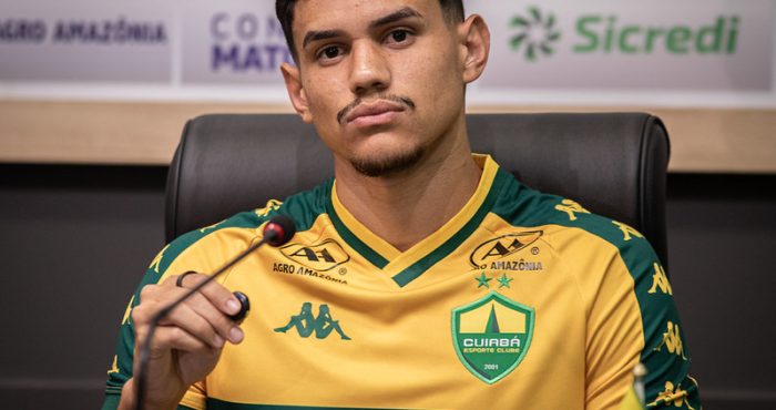 Close-up frontal de Edson sentado em uma cadeira de couro preta, posicionado atrás de um microfone durante uma coletiva de imprensa.