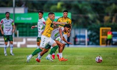 Uma imagem capturada em plano médio mostra uma partida de futebol profissional em um campo gramado durante o dia. No centro da ação, um jogador do Cuiabá (vestindo uniforme amarelo com detalhes verdes e calções brancos) disputa a posse de bola com um jogador do Juventude (vestindo uniforme branco com detalhes verdes).
