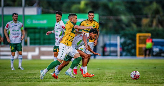 Uma imagem capturada em plano médio mostra uma partida de futebol profissional em um campo gramado durante o dia. No centro da ação, um jogador do Cuiabá (vestindo uniforme amarelo com detalhes verdes e calções brancos) disputa a posse de bola com um jogador do Juventude (vestindo uniforme branco com detalhes verdes).