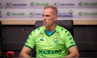 Goleiro Joao Carlos vestindo uma camisa de treino verde neon do Cuiabá Esporte Clube, sentado em uma coletiva de imprensa. Ele olha atentamente para o lado. Ao fundo, um painel com logotipos dos patrocinadores Sicredi e Agro Amazônia.
