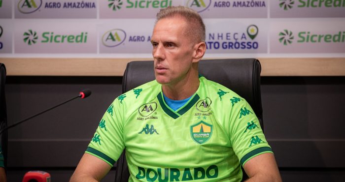 Goleiro Joao Carlos vestindo uma camisa de treino verde neon do Cuiabá Esporte Clube, sentado em uma coletiva de imprensa. Ele olha atentamente para o lado. Ao fundo, um painel com logotipos dos patrocinadores Sicredi e Agro Amazônia.