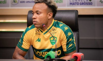 Mateus Santos sentado em uma mesa de coletiva de imprensa. Ele tem pele negra, cabelo curto com o topo descolorido e tatuagens no braço. Veste a camisa oficial de jogo do Cuiabá (amarela com detalhes verdes). À sua frente, microfones de rádio e TV.