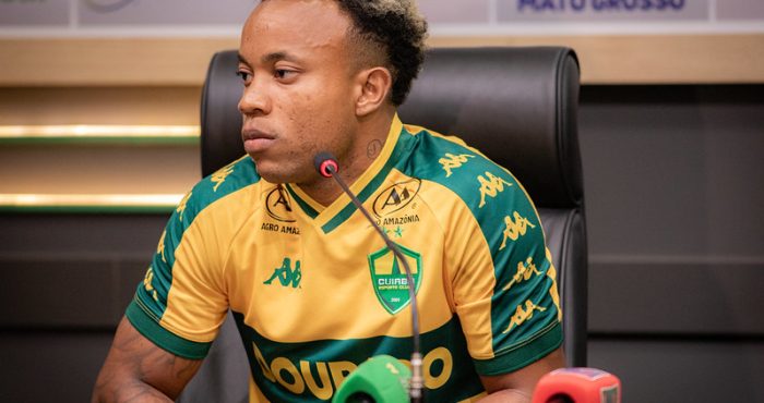 Mateus Santos sentado em uma mesa de coletiva de imprensa. Ele tem pele negra, cabelo curto com o topo descolorido e tatuagens no braço. Veste a camisa oficial de jogo do Cuiabá (amarela com detalhes verdes). À sua frente, microfones de rádio e TV.