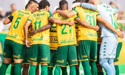 Jogadores do Cuiabá reunidos em um círculo apertado no campo, de costas para a câmera, abraçados pelos ombros em um momento de união antes ou depois de uma partida. É possível ver os nomes e números nas camisas amarelas, como Calebe (5), Eliel (11) e Gabriel (21), além do goleiro em seu uniforme azul à direita.
