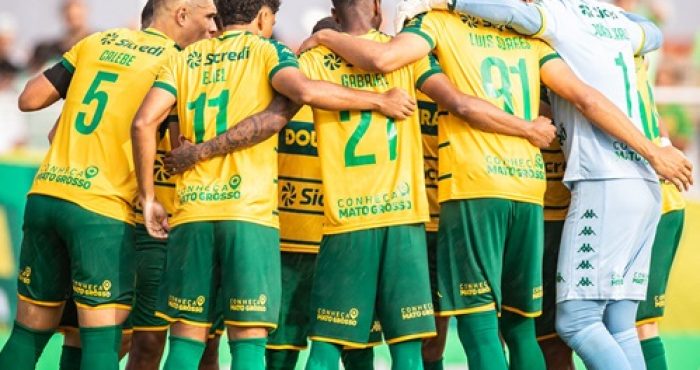 Jogadores do Cuiabá reunidos em um círculo apertado no campo, de costas para a câmera, abraçados pelos ombros em um momento de união antes ou depois de uma partida. É possível ver os nomes e números nas camisas amarelas, como Calebe (5), Eliel (11) e Gabriel (21), além do goleiro em seu uniforme azul à direita.