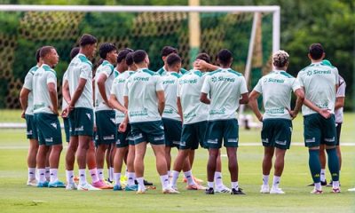 Fotografia em plano médio do grupo de jogadores do Cuiabá reunidos em um círculo no gramado durante um treinamento. Eles estão de costas para a câmera, vestindo uniformes de treino compostos por camisetas verde-claro com o patrocínio da Sicredi e calções verde-escuros. Ao fundo, vê-se uma trave de futebol com rede e uma área de vegetação densa sob luz natural.