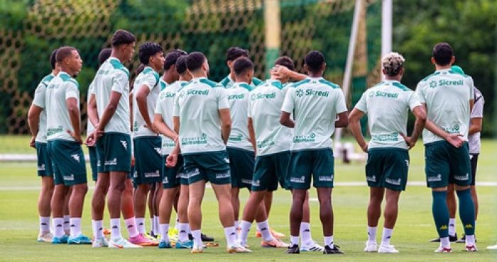 Fotografia em plano médio do grupo de jogadores do Cuiabá reunidos em um círculo no gramado durante um treinamento. Eles estão de costas para a câmera, vestindo uniformes de treino compostos por camisetas verde-claro com o patrocínio da Sicredi e calções verde-escuros. Ao fundo, vê-se uma trave de futebol com rede e uma área de vegetação densa sob luz natural.