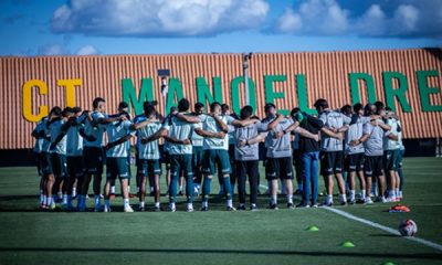 Mostra o grupo de jogadores e a comissão técnica reunidos em um círculo (o tradicional "huddle" ou roda de conversa) no gramado. Ao fundo, destaca-se o muro do centro de treinamento com a inscrição "CT MANOEL DRE..." (referindo-se ao CT Manoel Dresch). O céu está aberto com algumas nuvens, indicando condições ideais para o treino.