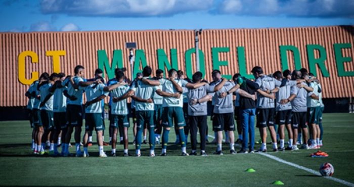 Mostra o grupo de jogadores e a comissão técnica reunidos em um círculo (o tradicional "huddle" ou roda de conversa) no gramado. Ao fundo, destaca-se o muro do centro de treinamento com a inscrição "CT MANOEL DRE..." (referindo-se ao CT Manoel Dresch). O céu está aberto com algumas nuvens, indicando condições ideais para o treino.
