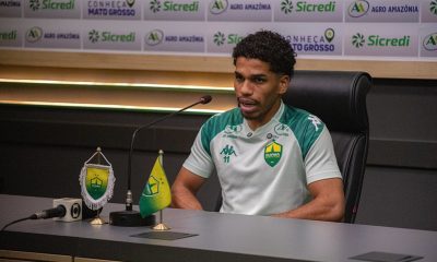 Eliel está sentado à frente de um painel de patrocinadores (backdrops), onde é possível ver marcas como Sicredi, Agro Amazônia e o selo "Conheça Mato Grosso". À sua frente, há microfones e um copo personalizado do clube.