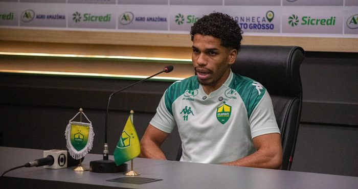 Eliel está sentado à frente de um painel de patrocinadores (backdrops), onde é possível ver marcas como Sicredi, Agro Amazônia e o selo "Conheça Mato Grosso". À sua frente, há microfones e um copo personalizado do clube.
