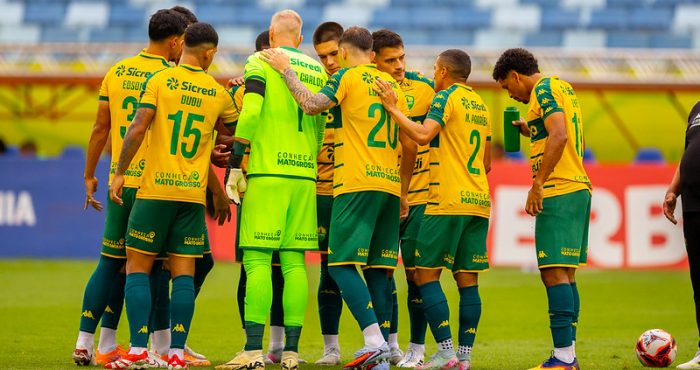 Jogadores do Cuiabá EC reunidos em círculo antes da partida na Arena Pantanal, usando uniforme amarelo e verde, em jogo do Campeonato Mato-grossense.