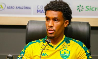 Close do jogador J. Araújo durante uma entrevista coletiva. Ele é um homem negro com cabelo crespo curto e cavanhaque, vestindo o uniforme de treino amarelo e verde do Cuiabá. Ele está sentado em uma cadeira de couro preta, falando ao microfone. O fundo mostra marcas de patrocinadores.