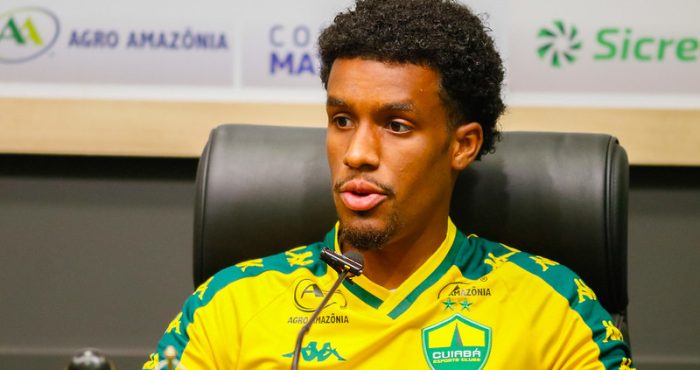 Close do jogador J. Araújo durante uma entrevista coletiva. Ele é um homem negro com cabelo crespo curto e cavanhaque, vestindo o uniforme de treino amarelo e verde do Cuiabá. Ele está sentado em uma cadeira de couro preta, falando ao microfone. O fundo mostra marcas de patrocinadores.