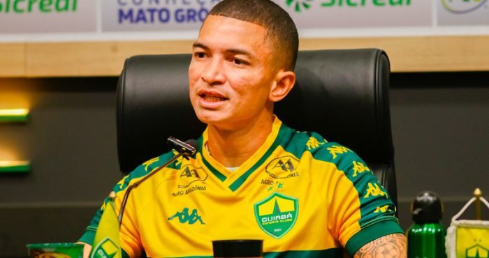 Close do jogador Marlon em uma coletiva de imprensa. Ele tem pele clara, cabelo raspado e uma tatuagem visível no braço esquerdo. Veste a camisa de treino amarela do clube e está posicionado em frente a um microfone, com uma expressão focada enquanto fala.