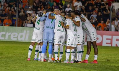 Elenco do Cuiabá Esporte Clube reunido em círculo no gramado antes da partida, jogadores abraçados em momento de concentração, com torcida ao fundo nas arquibancadas do estádio iluminado.