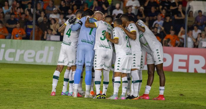 Elenco do Cuiabá Esporte Clube reunido em círculo no gramado antes da partida, jogadores abraçados em momento de concentração, com torcida ao fundo nas arquibancadas do estádio iluminado.
