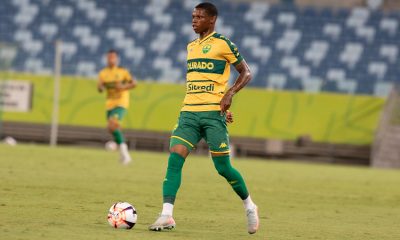 Jogador do Cuiabá Esporte Clube conduz a bola em partida oficial, vestindo uniforme amarelo e verde no gramado do estádio.