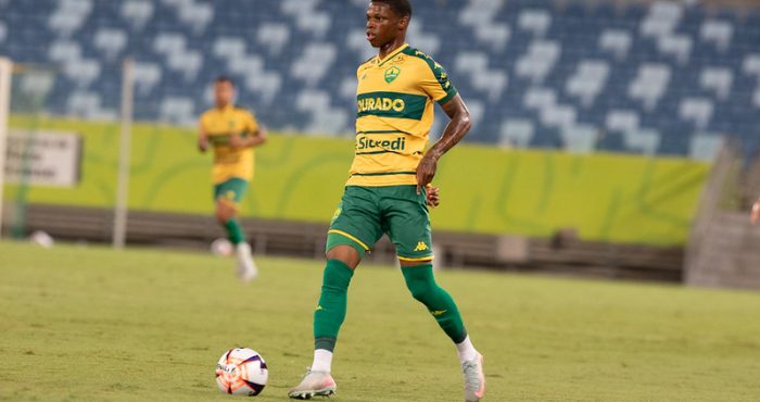 Jogador do Cuiabá Esporte Clube conduz a bola em partida oficial, vestindo uniforme amarelo e verde no gramado do estádio.