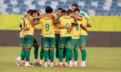 Foto do elenco do Cuiabá reunido em um círculo no centro do gramado. Os jogadores estão de costas, abraçados pelos ombros em um momento de união antes ou depois da partida. É possível ver os números 7, 9, 8 e 10 nos uniformes amarelos, além do goleiro à direita vestindo uniforme verde claro. Ao fundo, as arquibancadas do estádio estão vazias e iluminadas pelos refletores.